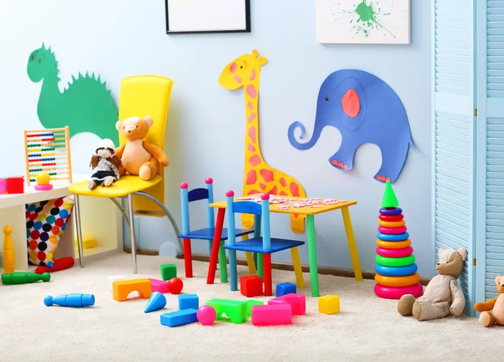 Una habitación de juegos colorida con decoraciones de animales en la pared, una silla con peluches, una mesa pequeña con sillas para niños y juguetes dispersos por el suelo.