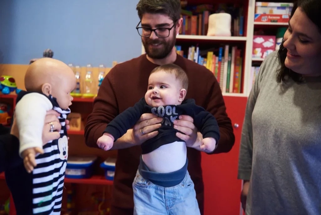 Un hombre y una mujer sonriendo mientras sostienen a dos bebés en una habitación llena de juguetes y libros.