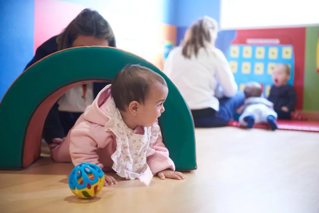 
Bebé gateando a través de un túnel de juego en una sala de estimulación temprana, con otros niños y educadores en el fondo.